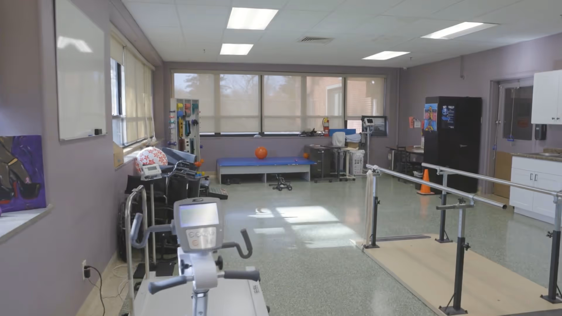 A physical therapy room with exercise equipment including a stationary bike, parallel bars for walking practice, a therapy table, and various therapy tools. The room has large windows with blinds, purple walls, and a linoleum floor.