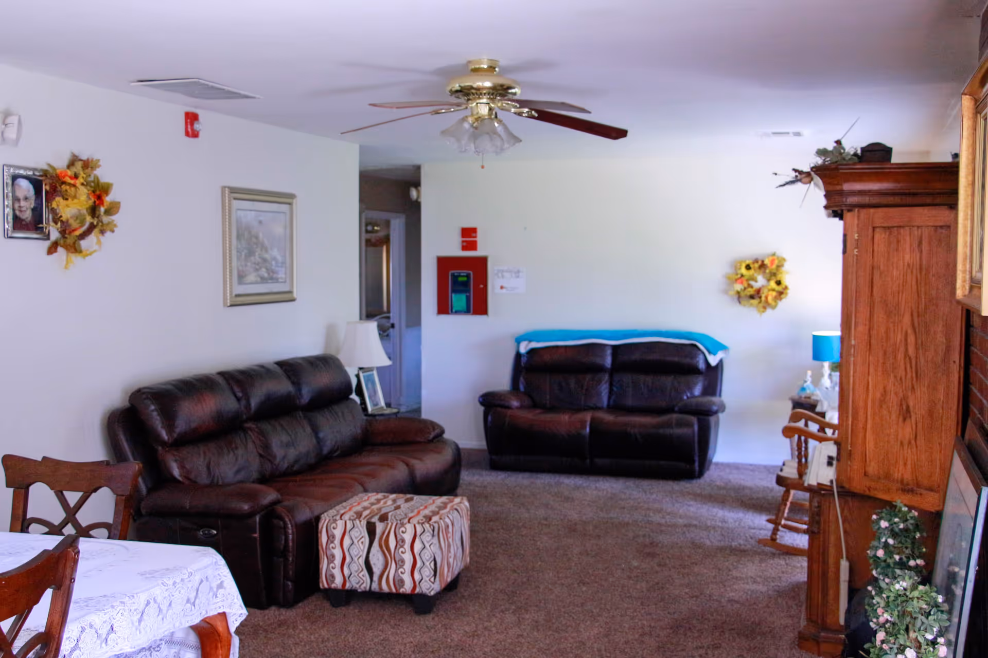 A cozy living room with two dark brown leather sofas, a patterned ottoman, a wooden rocking chair, and a wooden cabinet. The room has a ceiling fan with lights, a table with a white lace tablecloth, and wall decorations including framed pictures and wreaths. The carpet is brown, and there is a blue lamp on a side table near the far sofa.