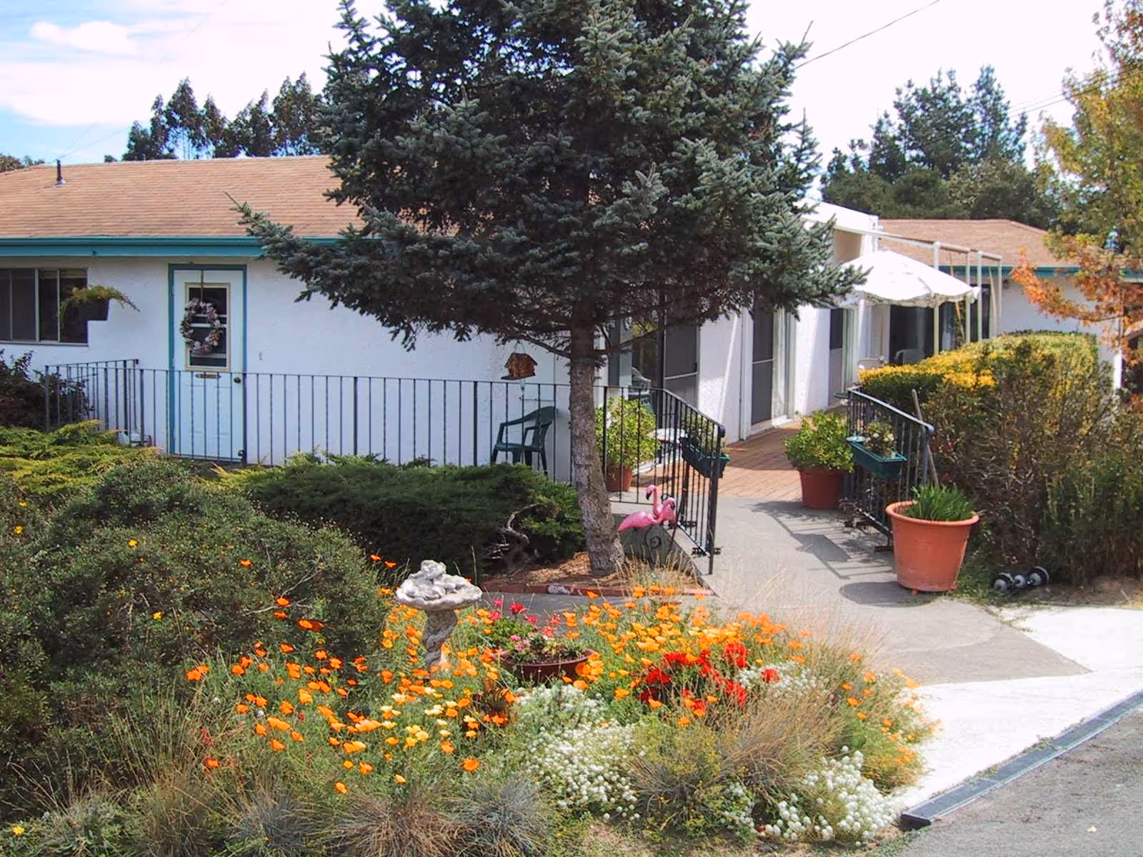 Outdoor view of a residential care facility with a garden featuring colorful flowers, shrubs, a small tree, and a birdbath. The building has a white exterior with a brown roof, a door with a wreath, and a walkway with railings leading to the entrance. There are potted plants and a pink flamingo garden decoration near the walkway.