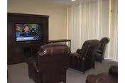 Small common living area with leather recliners facing a large TV in a wooden entertainment center and vertical blinds covering windows.