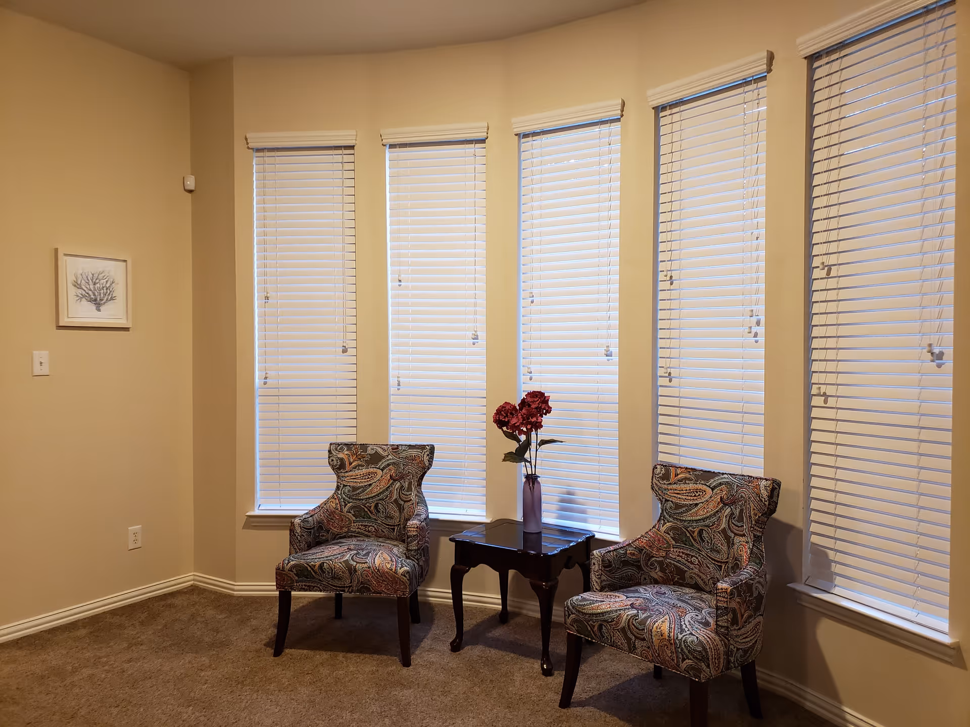 A cozy corner of a room with beige walls and carpeted floor, featuring two patterned armchairs with paisley designs placed on either side of a small dark wooden table holding a vase with red flowers. Behind the chairs are five tall windows with white blinds partially closed. A framed artwork hangs on the left wall.