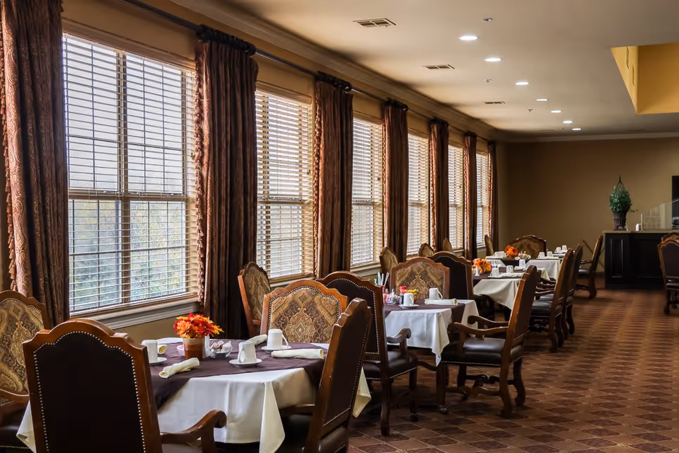 A dining room with multiple tables covered in white tablecloths and brown runners, each set with cups, napkins, and small flower arrangements. The room has large windows with wooden blinds and brown curtains, and the floor is tiled with a patterned design. The lighting is warm and recessed in the ceiling.