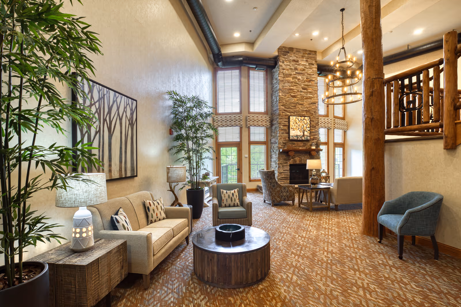 A spacious and warmly decorated living room area with high ceilings, large windows, and a stone fireplace. The room features comfortable seating including sofas and armchairs, wooden tables, potted plants, and a large chandelier. The decor includes natural wood elements and a patterned carpet.