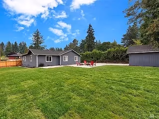 A spacious green lawn with a gray single-story building and a smaller gray shed. There is a concrete patio area with red chairs and a table. The sky is blue with scattered clouds, and there are tall trees and a wooden fence in the background.