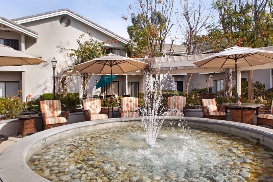 Outdoor courtyard area at a senior living facility with a circular water fountain in the center surrounded by cushioned wicker chairs and tables with large beige umbrellas. Trees and shrubs are planted around the seating area, and a building with windows and doors is visible in the background.