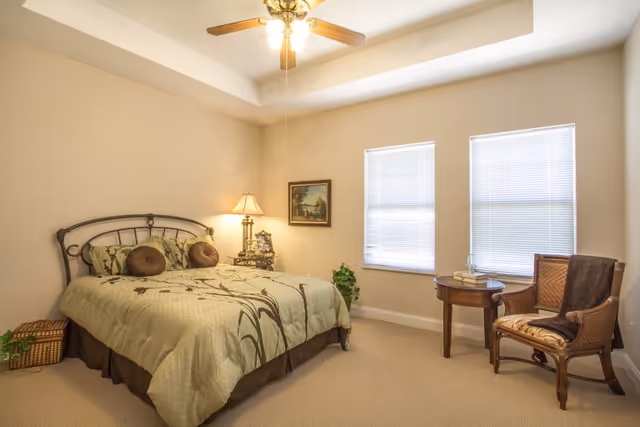 A cozy bedroom with a large bed featuring a green and brown floral comforter and two round pillows. There is a nightstand with a lamp and decorative items next to the bed. Two windows with blinds allow natural light into the room. A wooden chair with a cushion and a small round table with books and a glass of water are positioned near the windows. The room has beige walls and carpet, and a ceiling fan with a light fixture is mounted above.