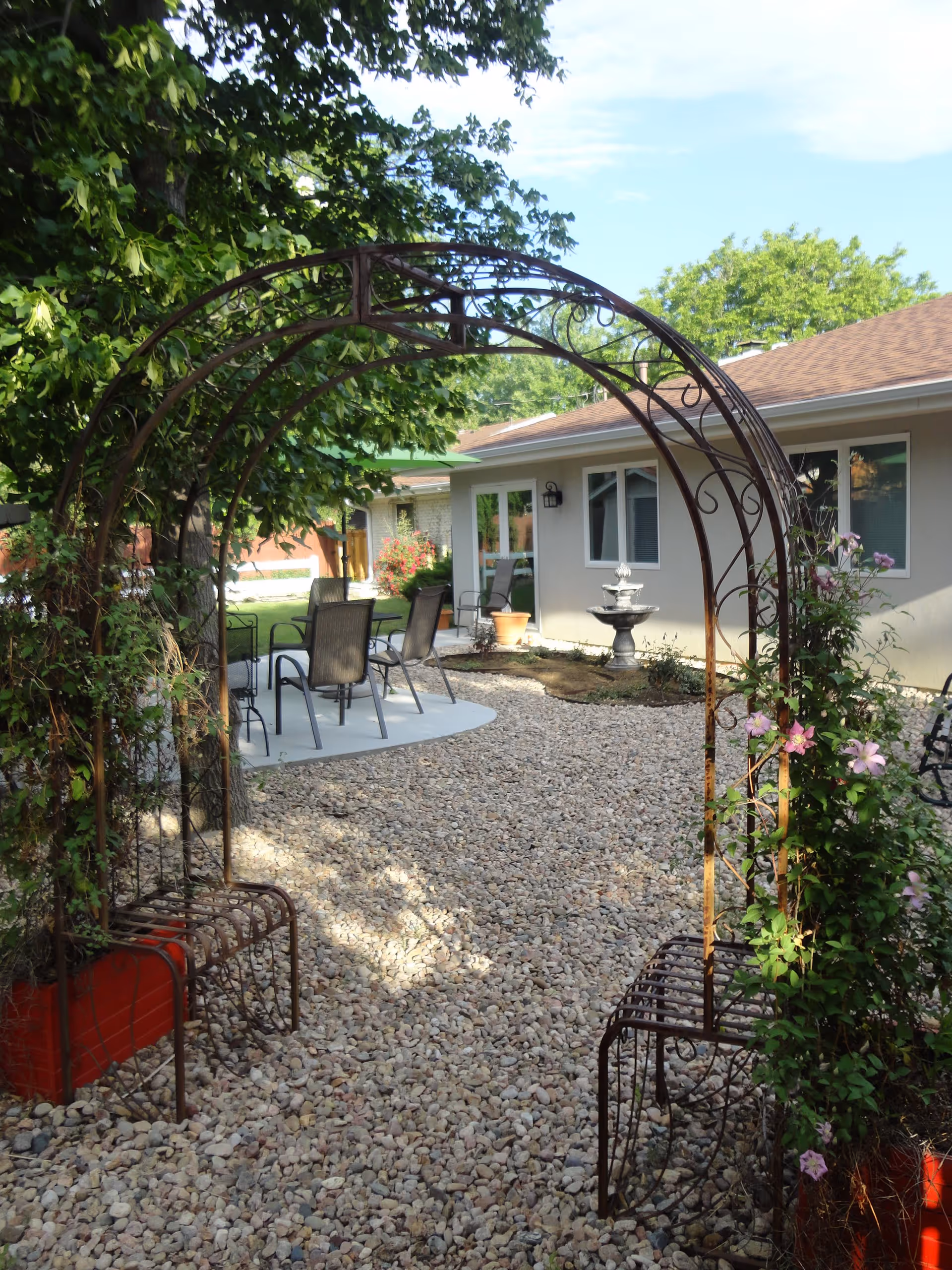 Outdoor garden area at Peak Gardens Assisted Living featuring a metal archway with climbing plants and flowers, gravel ground, patio chairs around a small concrete seating area, and a building with windows and a water fountain in the background.