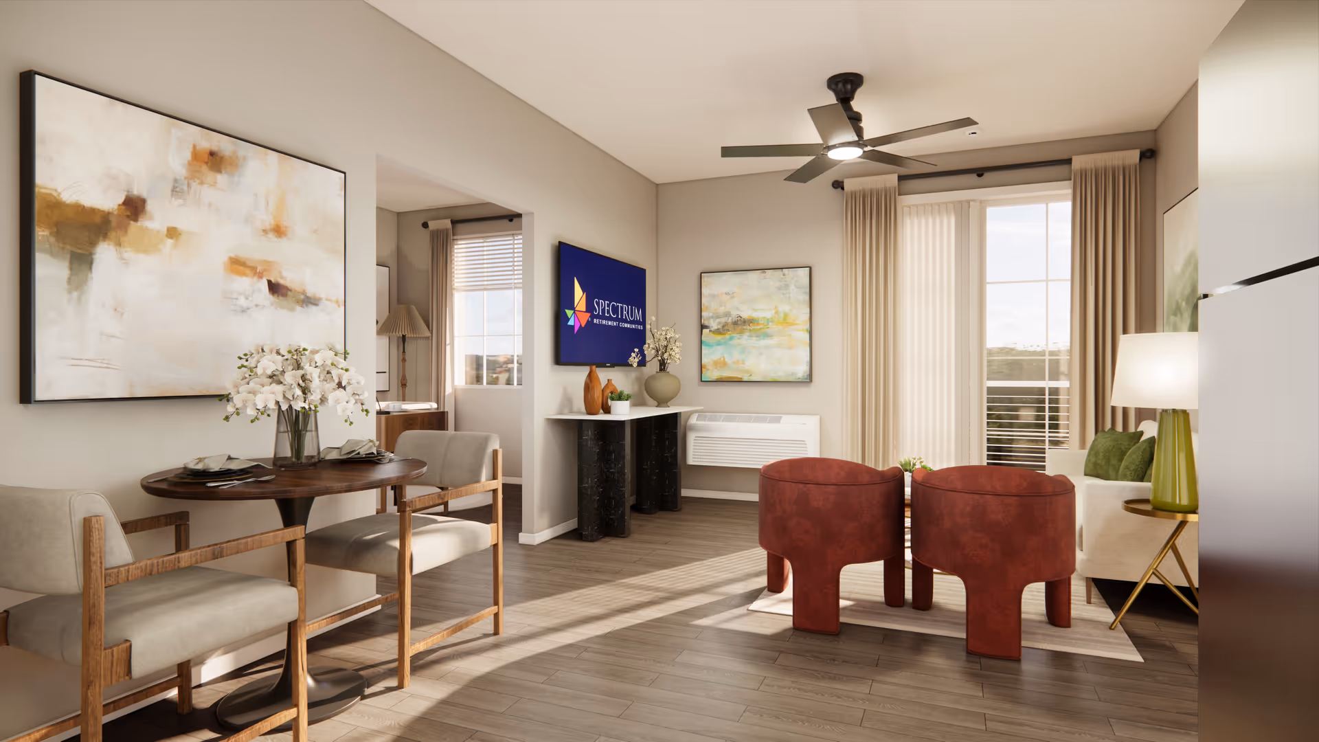 A bright and modern living room area in a senior living facility featuring a small round dining table with two chairs on the left, a wall-mounted TV displaying the Spectrum Retirement Communities logo, two red armchairs facing a sofa with green cushions, a floor lamp with a green base, abstract paintings on the walls, large windows with beige curtains, and a ceiling fan.