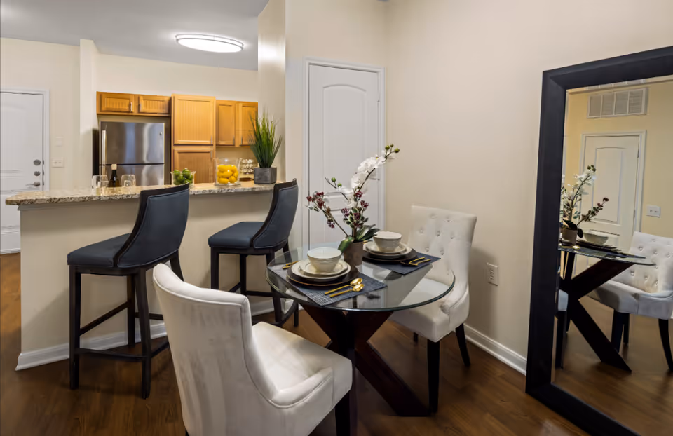 A modern dining area with a round glass table set for two with white cushioned chairs. Behind the table is a kitchen counter with two dark upholstered bar stools. The kitchen features wooden cabinets and a stainless steel refrigerator. A large black-framed mirror is on the wall reflecting part of the room.