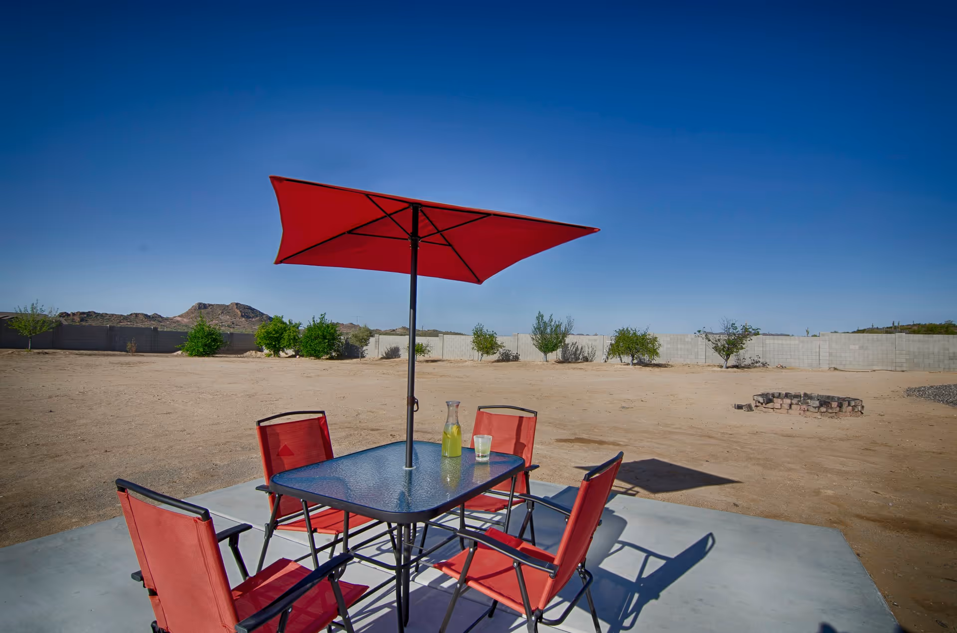 Outdoor patio area with a glass table, four red chairs, and a large red umbrella providing shade. The setting is in a desert-like environment with sparse vegetation and a clear blue sky.