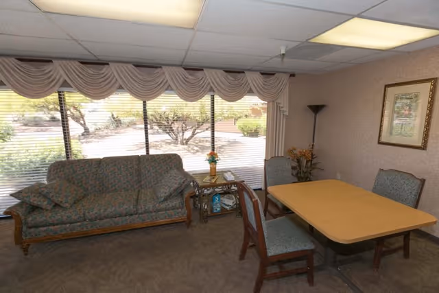 A small common room with a patterned sofa, a table with chairs and large windows with draped valances looking out to landscaping.