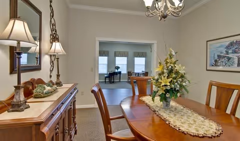 A dining room with a wooden table and chairs, a floral centerpiece on the table, a sideboard with decorative items and lamps, and a view into an adjacent room with a piano and windows.