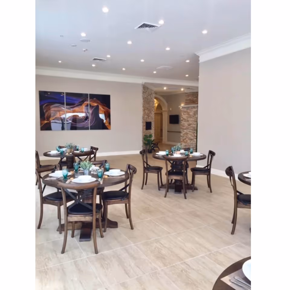 A bright dining room with round wooden tables set with white plates, teal glasses, and small green plants as centerpieces. The room has light-colored tiled floors, beige walls, recessed ceiling lights, and a modern abstract painting on one wall. There is a stone accent wall and an arched doorway in the background.
