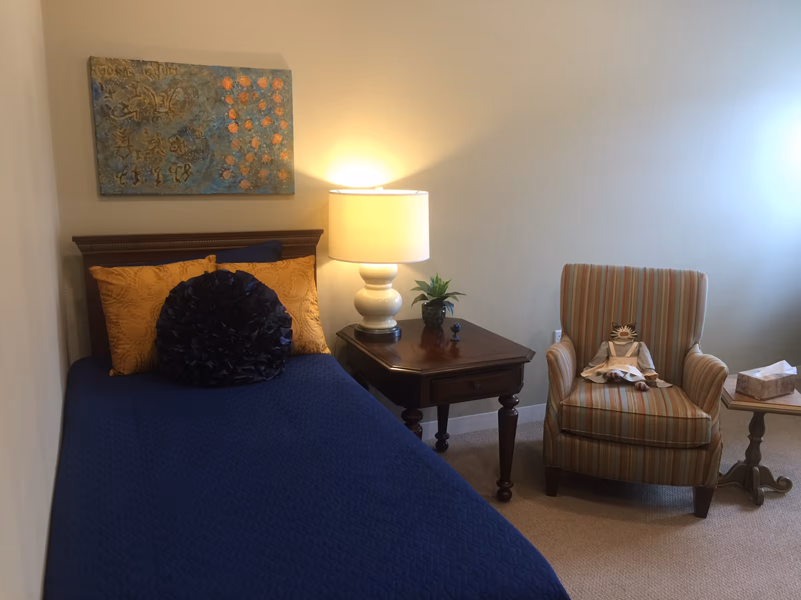 A cozy bedroom with a single bed covered in a blue bedspread and decorated with two yellow pillows and a round dark blue pillow. Next to the bed is a wooden nightstand with a white lamp and a small green plant. On the right side, there is a striped armchair with a doll sitting on it, and a small wooden side table with a tissue box. A piece of abstract artwork hangs on the wall above the bed.