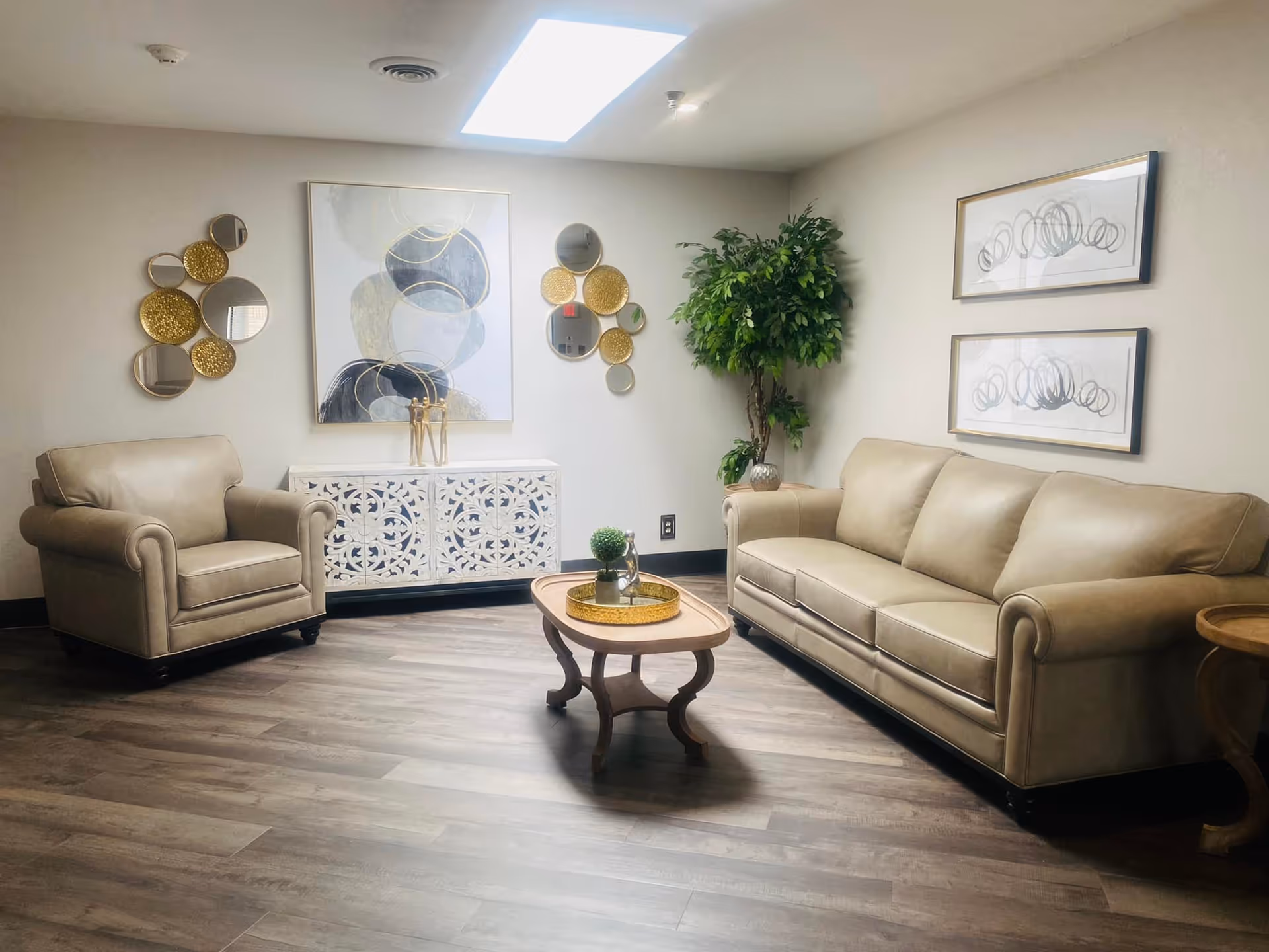 A cozy living room area with a beige leather armchair and sofa, a wooden coffee table with decorative items, a white ornate cabinet against the wall, abstract wall art, circular gold and mirror wall decorations, and a tall green potted plant in the corner. The room has wooden flooring and a skylight providing natural light.