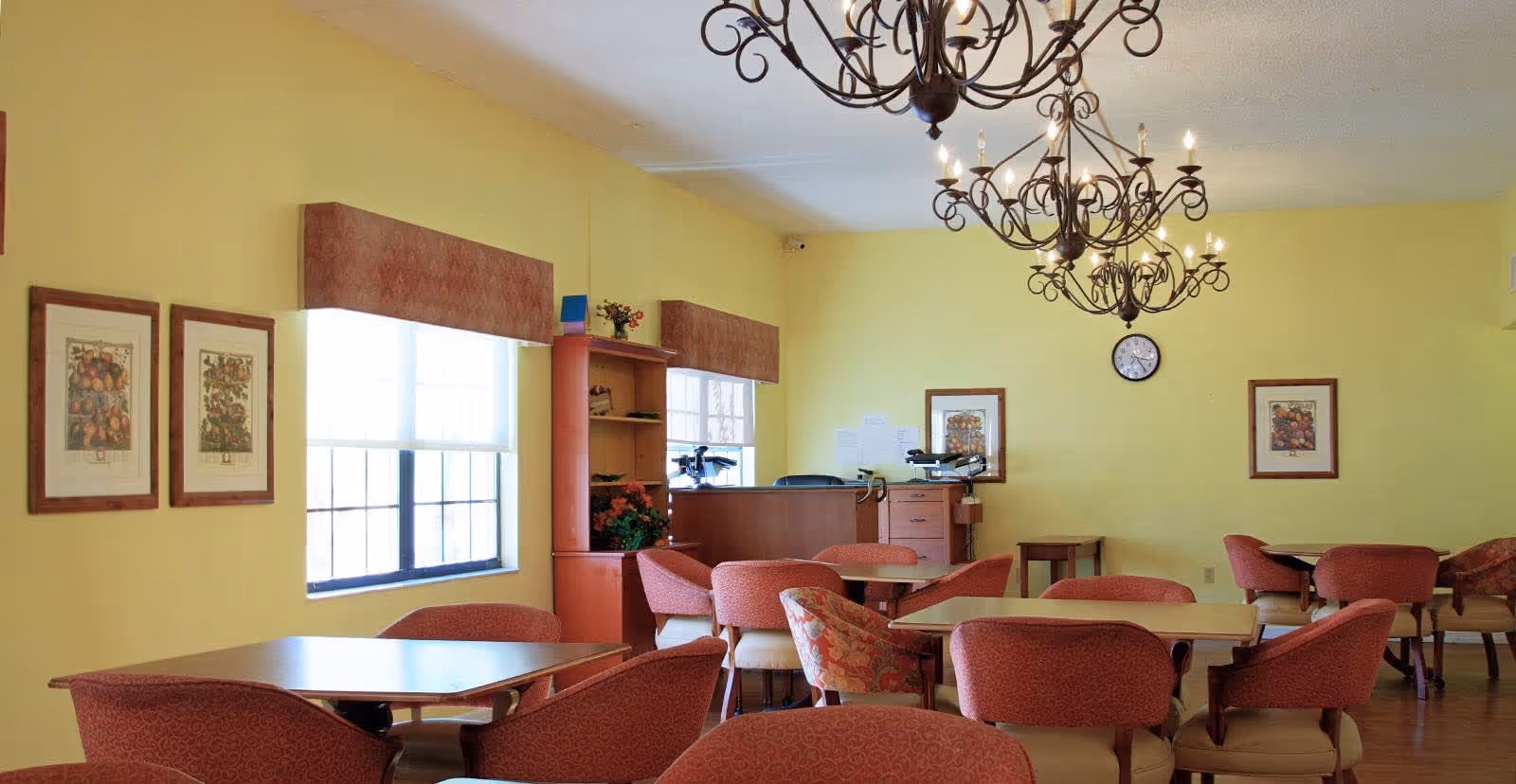 A dining room with yellow walls, several tables surrounded by red cushioned chairs, two large windows with valances, framed artwork on the walls, a clock, and two ornate chandeliers hanging from the ceiling.