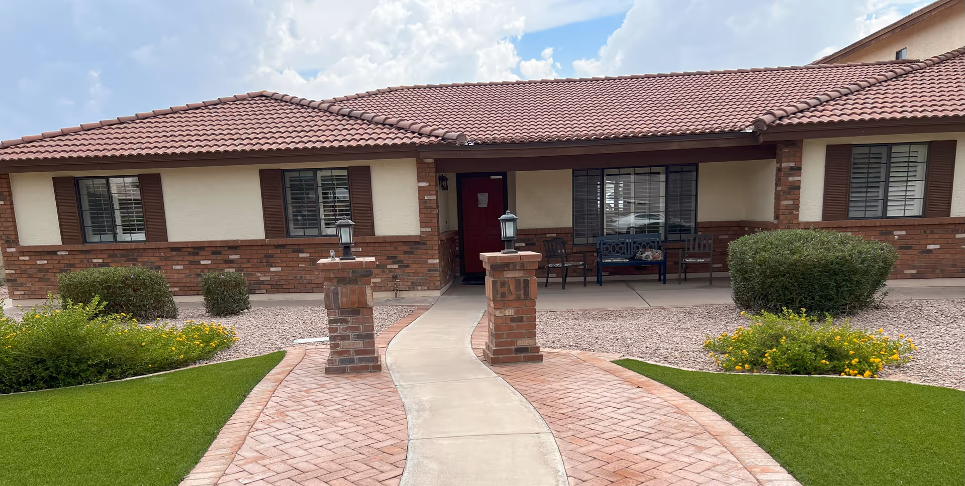 Front exterior view of a single-story brick and stucco building with a red tiled roof, a curved concrete walkway leading to a red front door, two brick pillars with lanterns, green bushes, yellow flowers, and a blue bench with chairs on the porch under a partly cloudy sky.