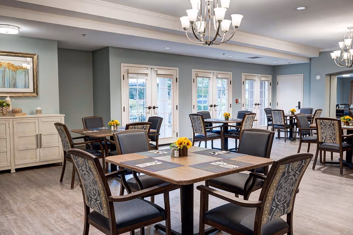 A bright and spacious dining room with multiple wooden tables and cushioned chairs arranged neatly. Each table is set with placemats, utensils, and small flower centerpieces. The room features light blue walls, large windows with French doors allowing natural light to enter, and elegant chandeliers hanging from the ceiling. A sideboard with decorative items and a framed painting is visible on one wall.