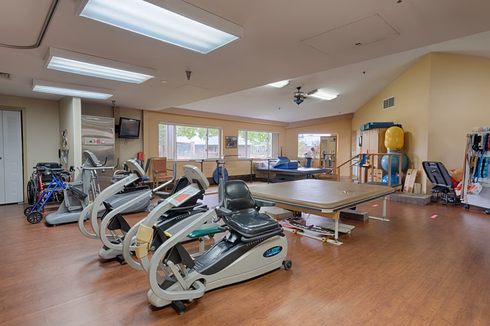 A spacious rehabilitation or physical therapy room with exercise equipment including stationary bikes, parallel bars, therapy tables, and various therapy balls. The room has large windows letting in natural light and wood flooring.