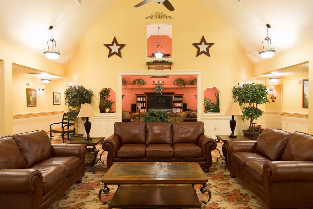 A cozy living room area with three brown leather sofas arranged around a wooden coffee table on a floral carpet. The walls are painted yellow with two large star decorations and a mirror above the central sofa. There are two side tables with lamps and potted plants on either side of the central sofa. The room is well-lit with ceiling lights and has an open doorway leading to another room with a television and more plants.