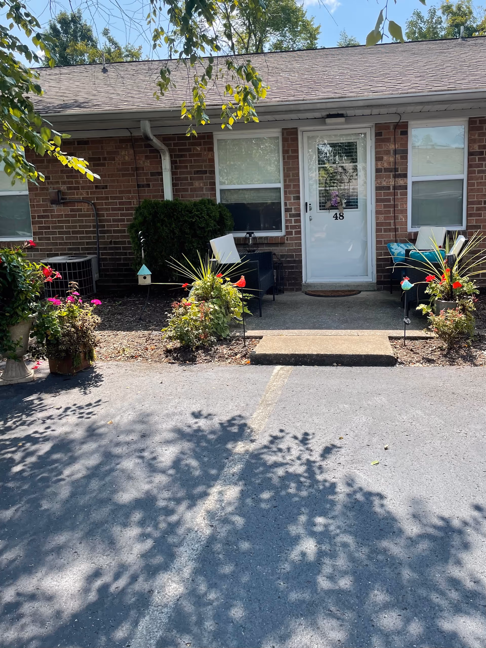 Exterior view of a single-story brick building with a white door numbered 48. There are two windows on either side of the door, outdoor chairs with cushions, and potted plants with flowers arranged near the entrance. The area is shaded by tree branches, and there is a paved parking area in front.