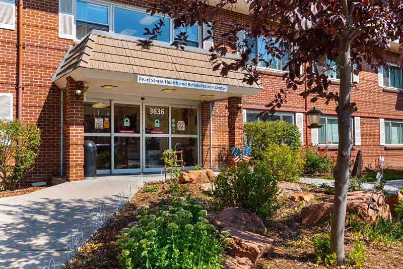 Entrance of a brick building with a sign above the door reading 'Pearl Street Health and Rehabilitation Center'. The building has large windows and a small garden with plants and rocks in front. A concrete pathway leads to the glass double doors with the number 3636 displayed above.