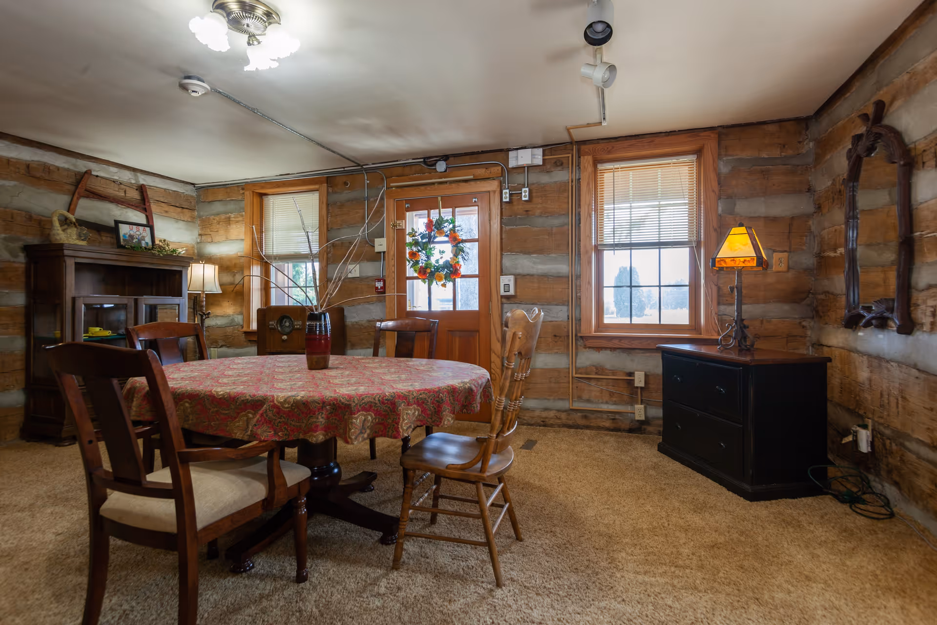 Interior of a rustic dining room with log cabin walls, a round table covered with a patterned tablecloth, four wooden chairs, a wooden cabinet with a lamp and decorative items, a door with a floral wreath, two windows with blinds, and a small black dresser with a lamp and a mirror above it.