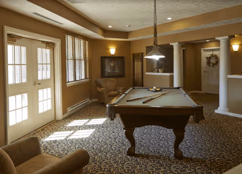 A cozy interior room with a pool table in the center, two pool cues resting on the table, a hanging light fixture above, patterned carpet, beige walls, a comfortable armchair, framed artwork on the wall, and French doors letting in natural light.