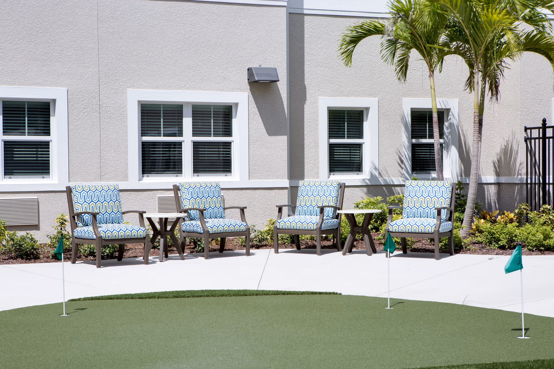 Outdoor courtyard with four patterned lounge chairs and small tables beside a putting green in front of a beige building with windows and palm trees.