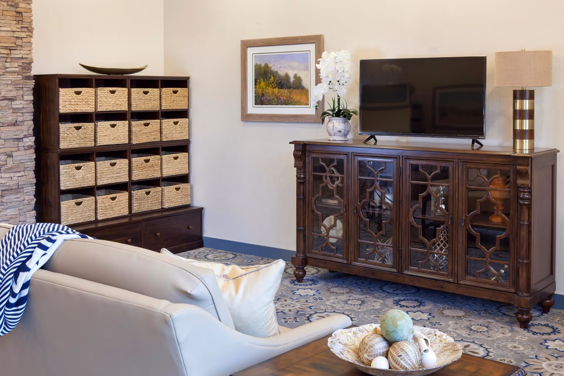 A bright living room with a TV on an ornate wooden console, wicker storage cubbies, a beige armchair, and decorative rug and accessories.