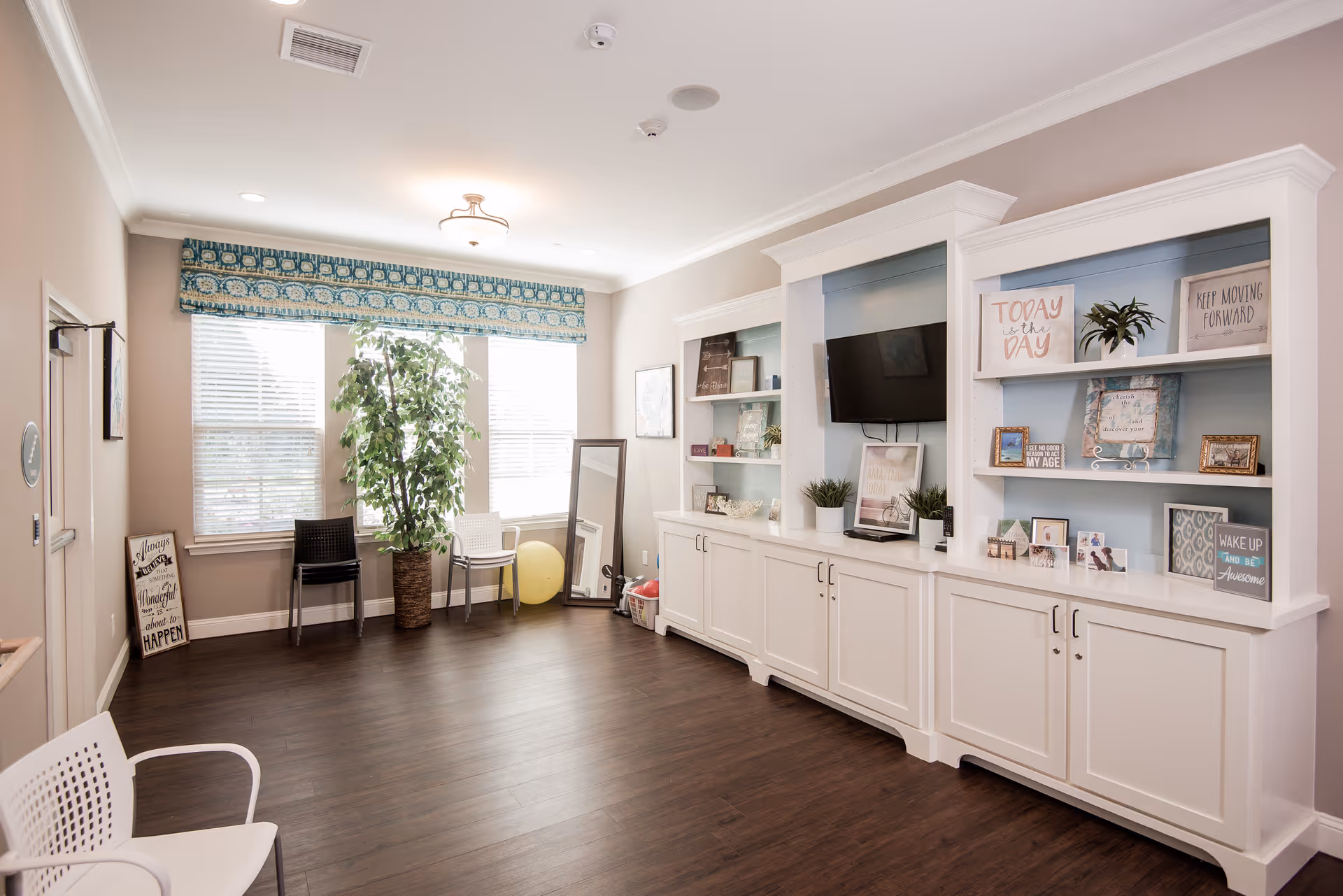 Bright communal living room with white built-in shelving and TV, seating, and large windows with a potted plant.