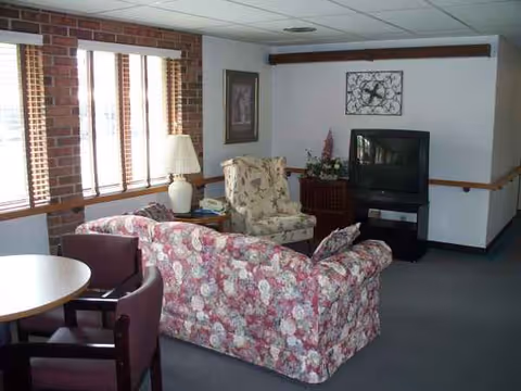 Cozy senior living lounge with floral sofa, chairs, lamp, windows, and television.