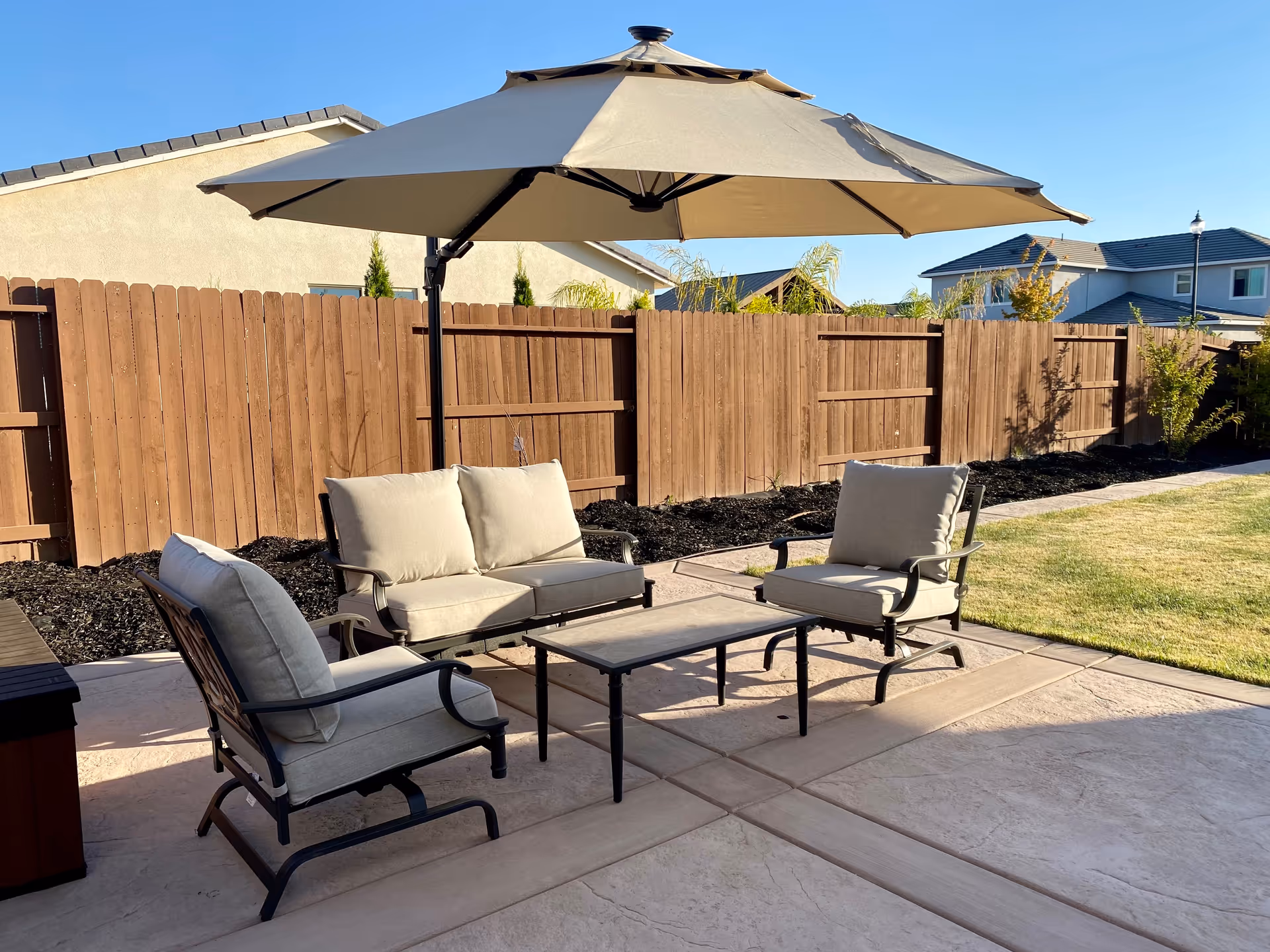 Outdoor patio area with cushioned seating including a loveseat and two chairs around a rectangular coffee table, shaded by a large beige umbrella. The patio is paved and bordered by a wooden fence with a grassy lawn adjacent to it. Residential houses are visible in the background under a clear blue sky.