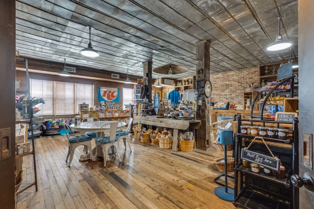 Rustic indoor retail-style common area with wooden floors, a round table and chairs, and shelves and displays of goods under hanging lights.