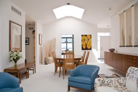 Bright open living and dining area with blue armchairs, a round wooden dining table, skylight, and wall artwork.