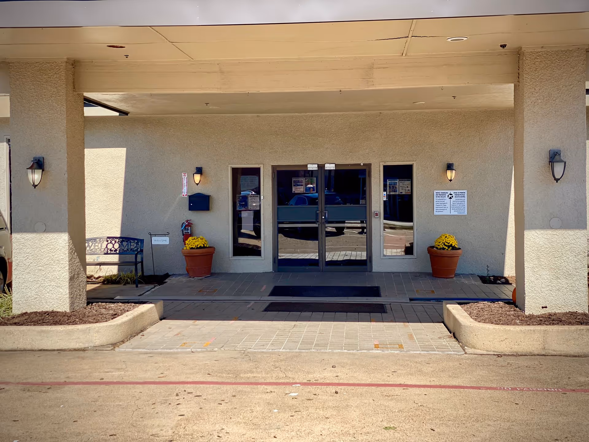 Covered main entrance of Terrell Healthcare Center with double glass doors, potted flowers, wall lamps and a bench.