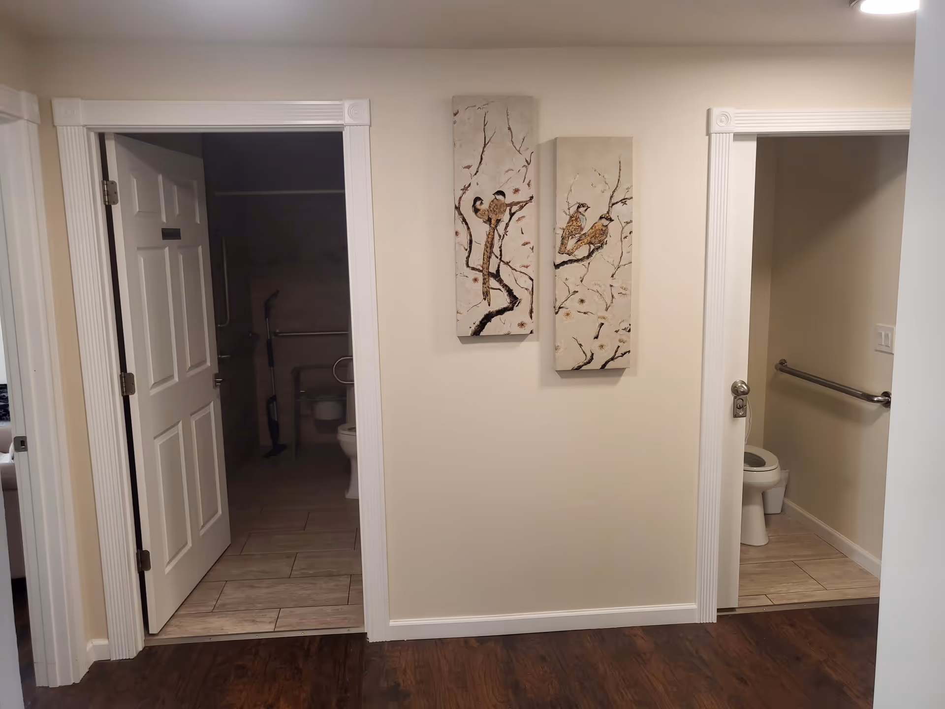 Interior view showing two open doorways leading to separate bathrooms with toilets and grab bars. Between the doorways, two vertical paintings of birds on branches hang on a beige wall. The floor outside the bathrooms is dark wood, while the bathroom floors are tiled.