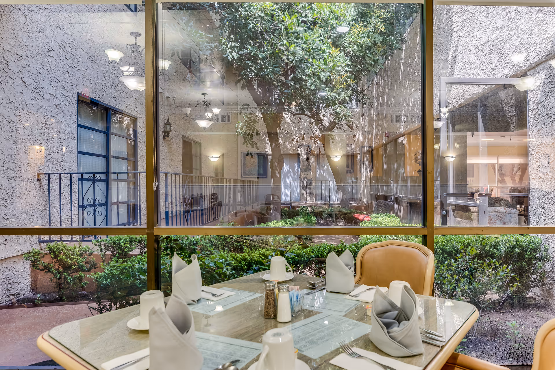 A dining table set for four with folded napkins, utensils, and condiments in a senior living facility. The table is positioned next to large glass windows that look out onto a small courtyard with green bushes and a tree. The courtyard is surrounded by the building's interior walls and has outdoor lighting fixtures.