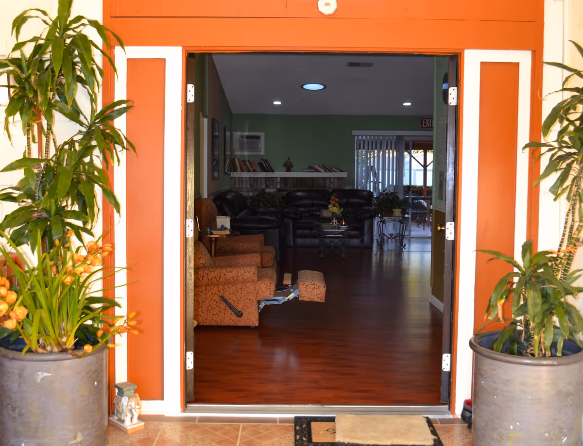 View through an open doorway into a living room with wooden flooring, featuring a brown patterned recliner chair, black leather sofas, a coffee table with a flower arrangement, and a bookshelf above a brick fireplace. Two large potted plants flank the doorway, which has orange and white trim.
