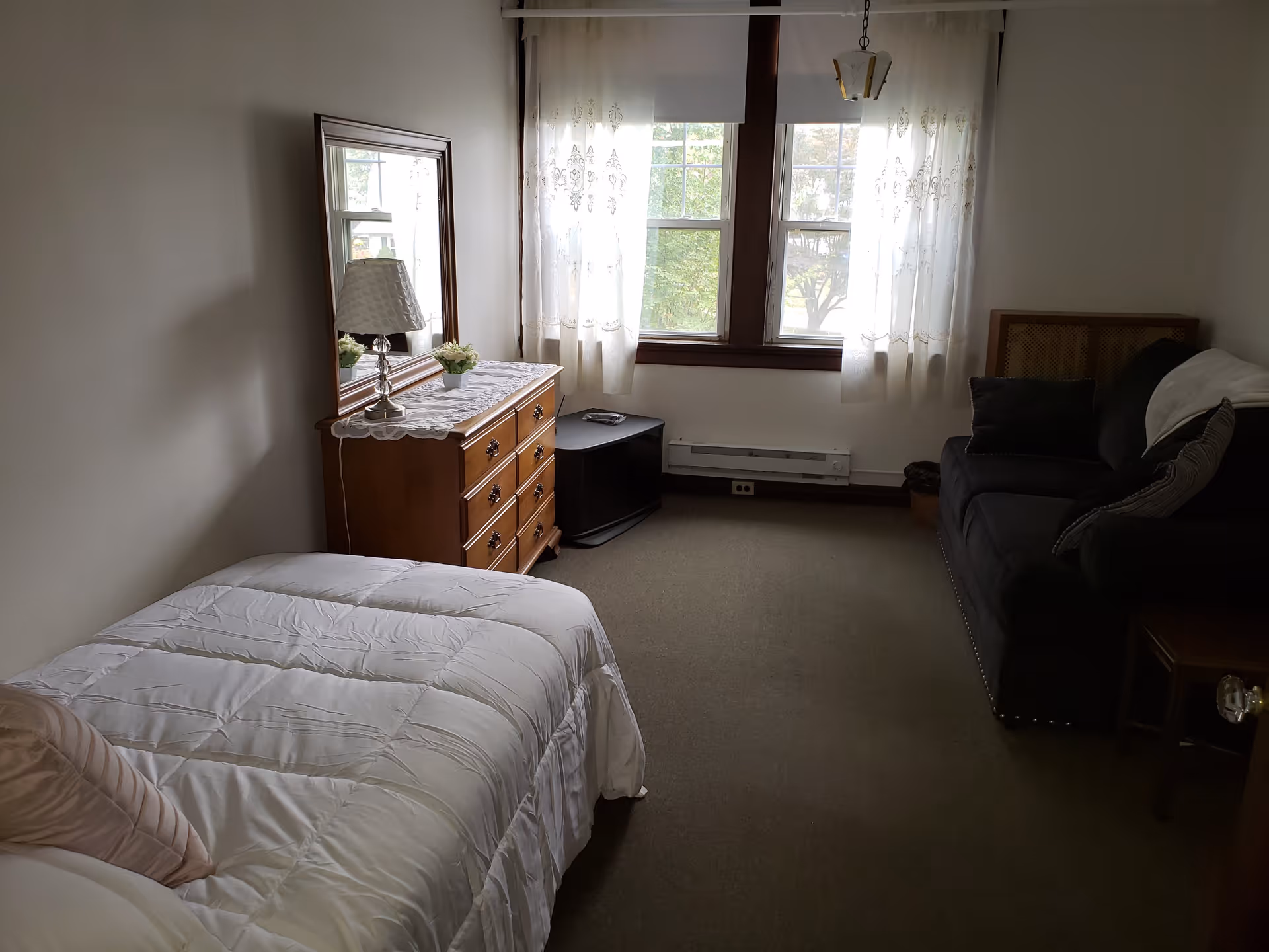 A cozy bedroom with a single bed covered in a white comforter and a beige pillow. Next to the bed is a wooden dresser with a lace runner, a table lamp, and a small potted plant. A large mirror is mounted on the wall above the dresser. Across from the bed is a dark-colored couch with cushions and a small side table. Two windows with sheer white curtains allow natural light into the room.