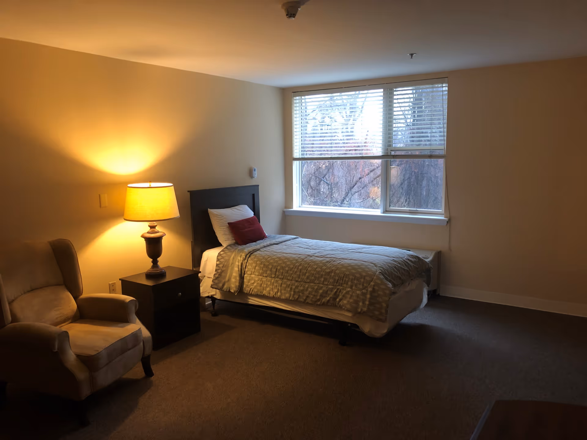 A simple bedroom with a single bed covered with a light gray patterned blanket and a red pillow. Next to the bed is a dark wooden nightstand with a lit table lamp. To the left of the nightstand is a beige upholstered armchair. A large window with white blinds lets in natural light, showing trees outside. The walls are painted a light beige color and the floor is carpeted.