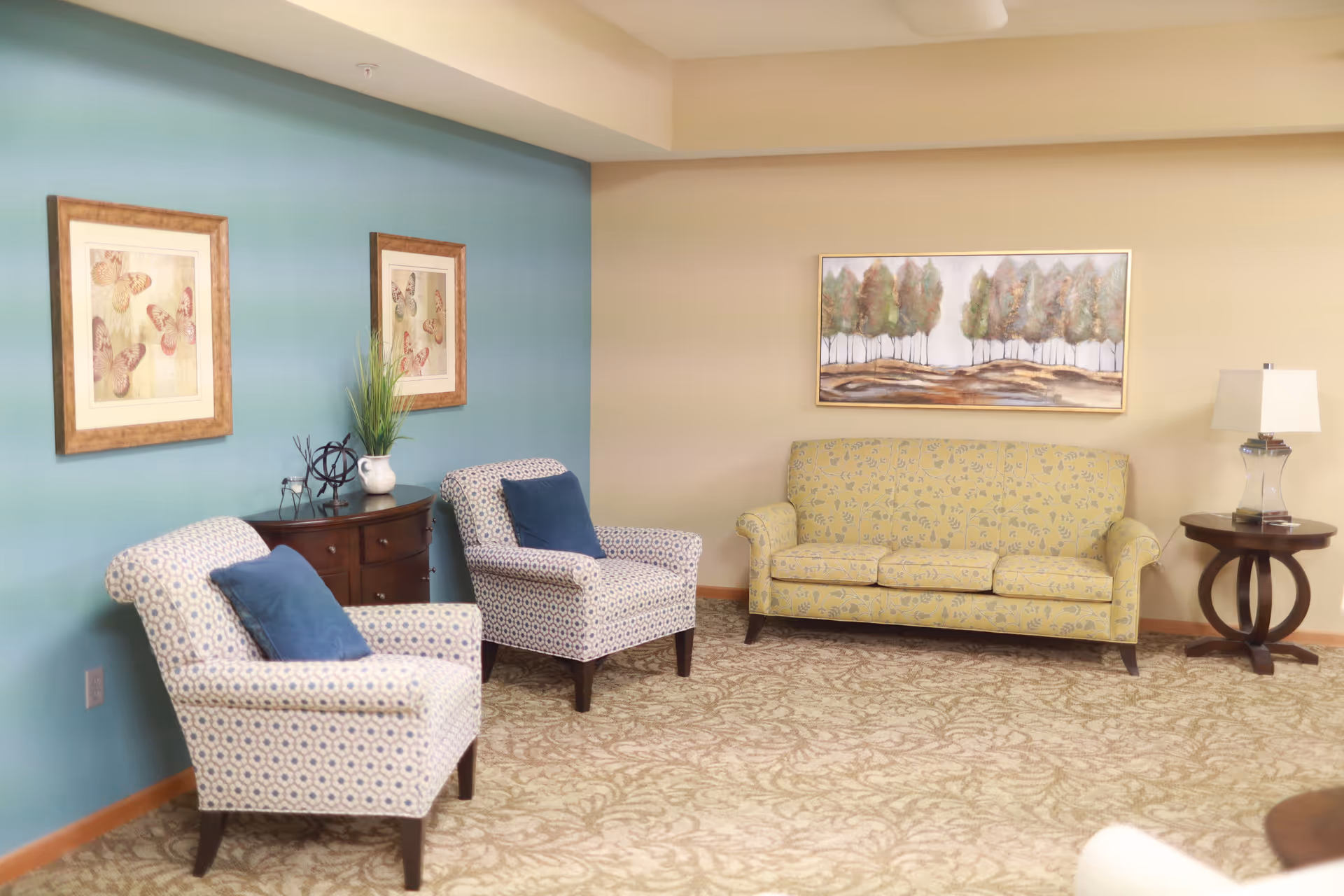 A cozy living room area with two patterned armchairs featuring blue cushions, a yellow floral-patterned sofa, a wooden side table with a lamp, and framed artwork on the walls. The room has a blue accent wall and a beige carpet with a subtle pattern.