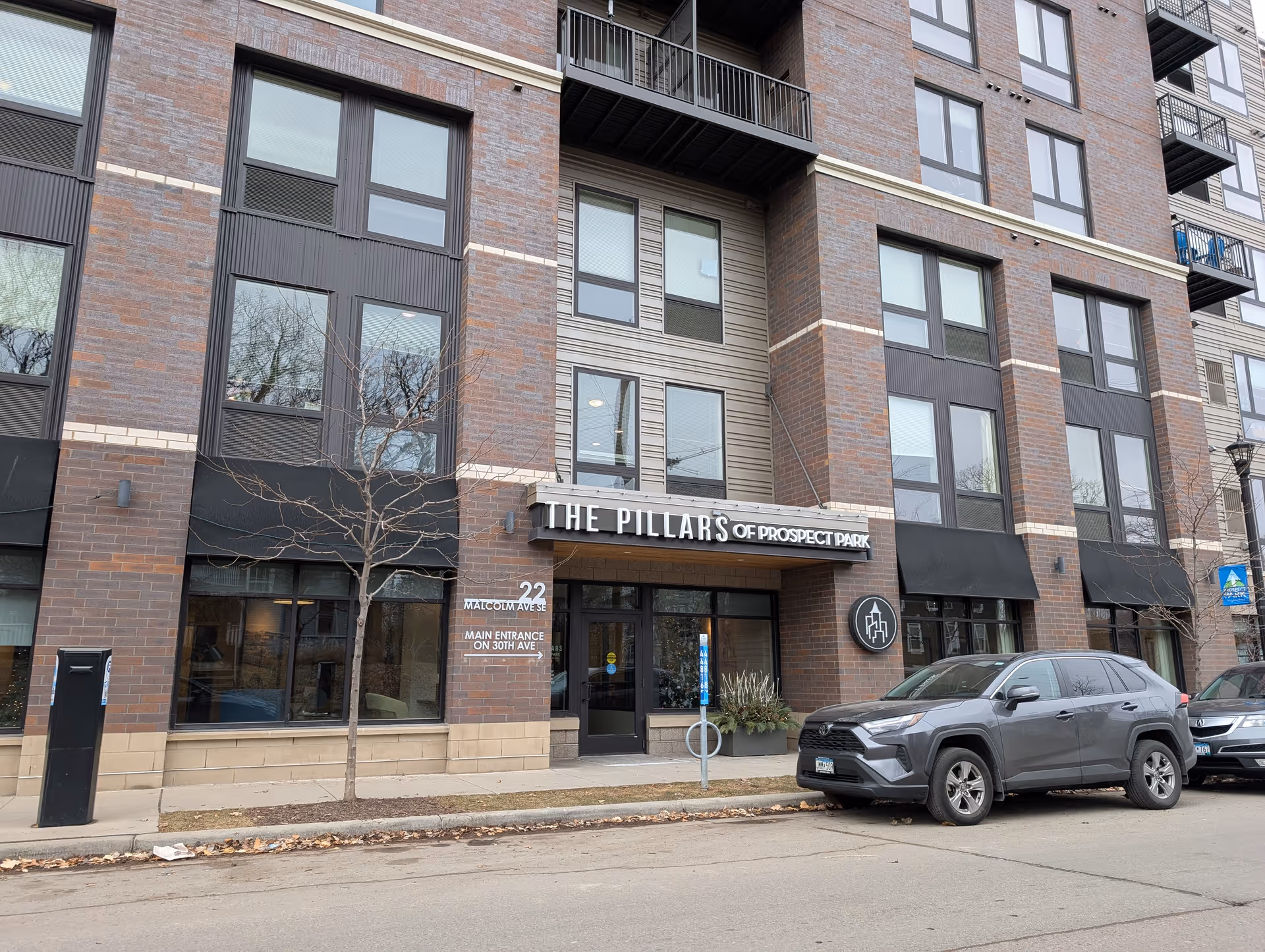 Exterior view of a modern multi-story brick building with large windows and a sign above the entrance that reads 'The Pillars of Prospect Park'. There are two leafless trees on the sidewalk and two parked cars in front of the building. The address '22 Malcolm Ave SE' and a note about the main entrance on 30th Ave are visible on the wall near the entrance.