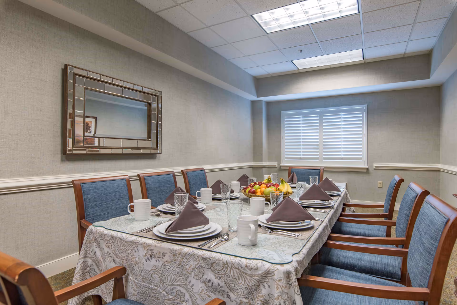 A dining room with a rectangular table set for eight people. The table is covered with a patterned tablecloth and a glass top, and each place setting includes a plate, a folded brown napkin, a glass, a coffee mug, and silverware. A centerpiece bowl filled with assorted fruits is in the middle of the table. The room has beige walls, a large mirror on one wall, a window with white shutters, and wooden chairs with blue upholstered seats and backs.