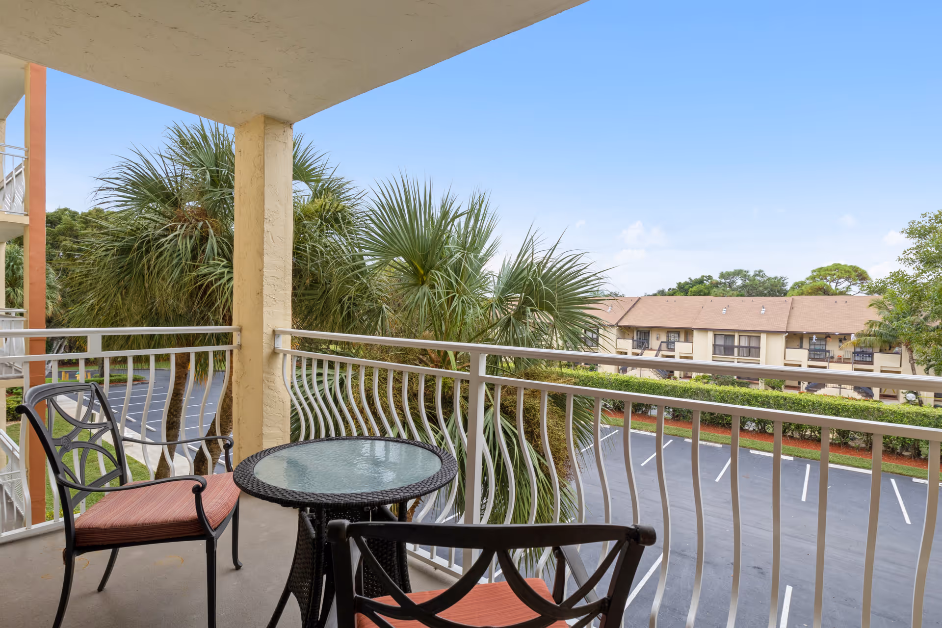 Covered balcony with a glass-top table and two chairs overlooking palm trees, a parking lot, and neighboring apartments.