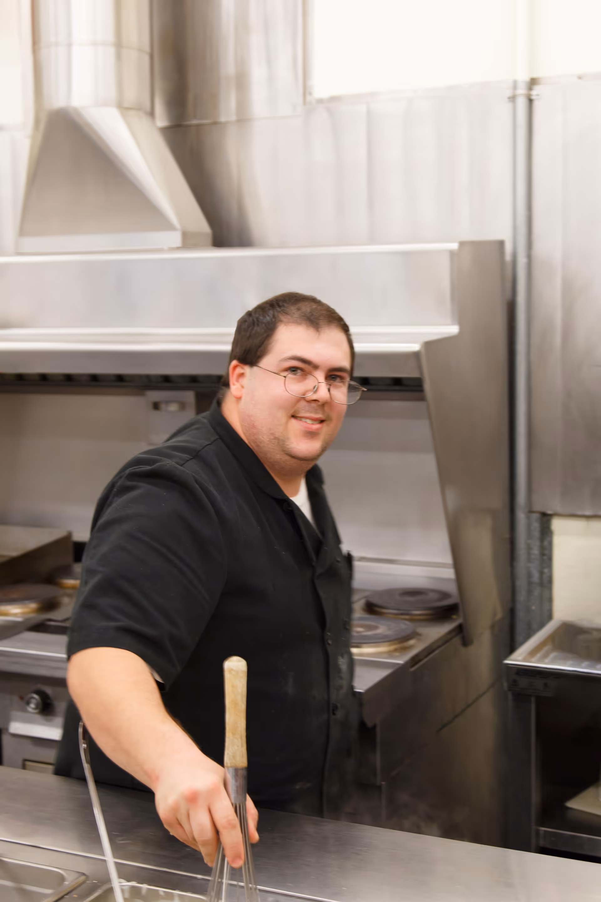 A man wearing glasses and a black chef coat is standing in a commercial kitchen, smiling at the camera while holding a whisk. Stainless steel kitchen appliances and a stove are visible in the background.