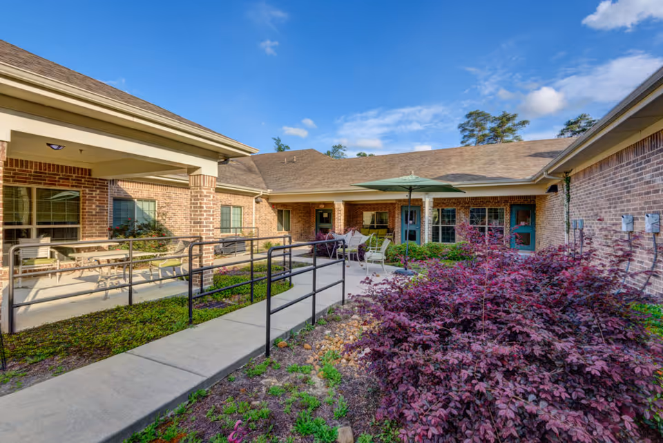 Outdoor courtyard area of a senior living facility with a concrete walkway and black metal handrails, surrounded by green and purple foliage. The building is made of brick with multiple windows and doors, and there is a patio area with chairs and a green umbrella under a clear blue sky.