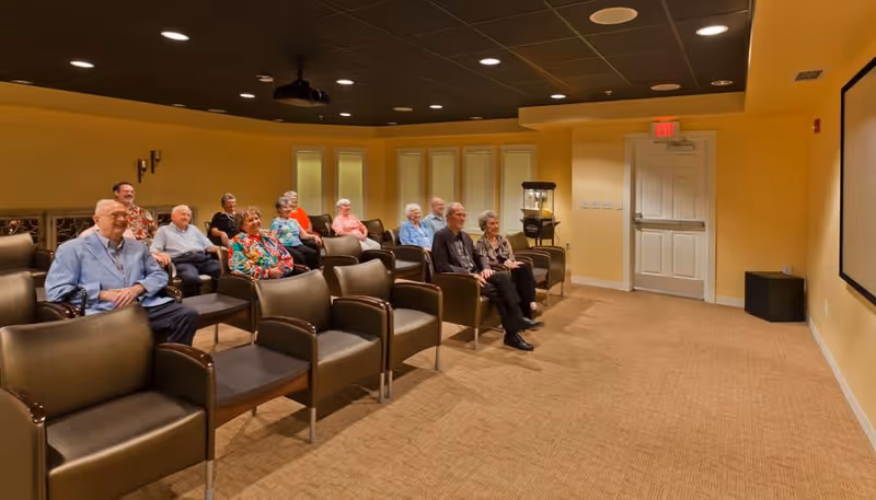 A group of elderly people seated in rows of brown chairs in a small theater or media room with a projector on the ceiling and a large screen on the wall. The room has yellow walls, a carpeted floor, and a popcorn machine near the back door.