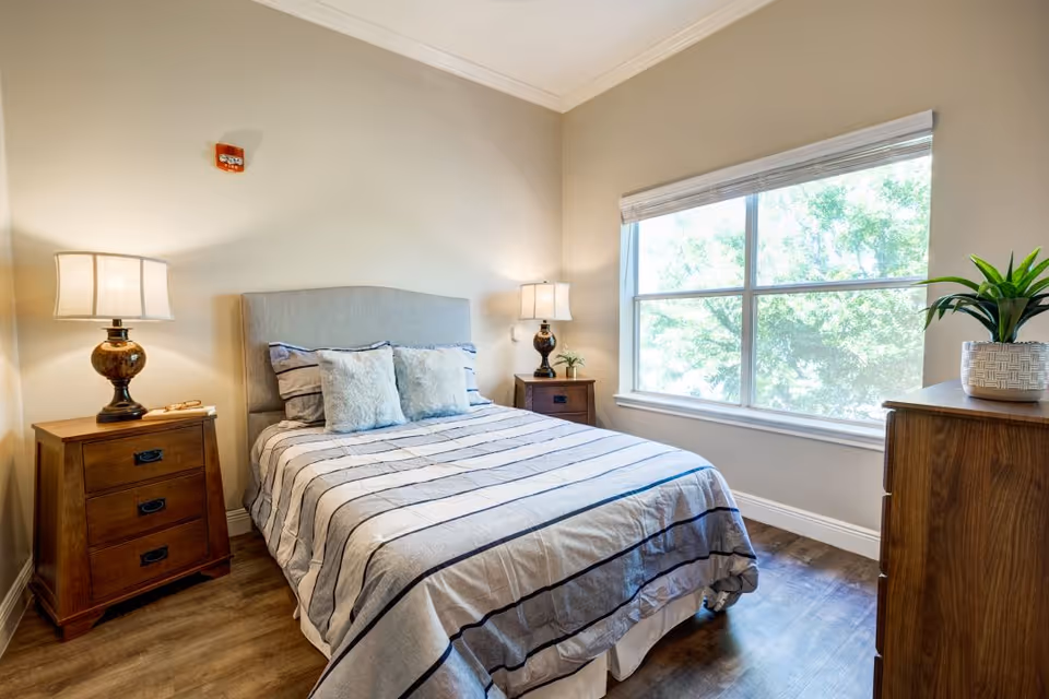 A bright bedroom with a large window showing green trees outside. The room features a bed with a striped bedspread and multiple pillows, flanked by two wooden nightstands each with a lamp. There is also a wooden dresser with a potted plant on top.