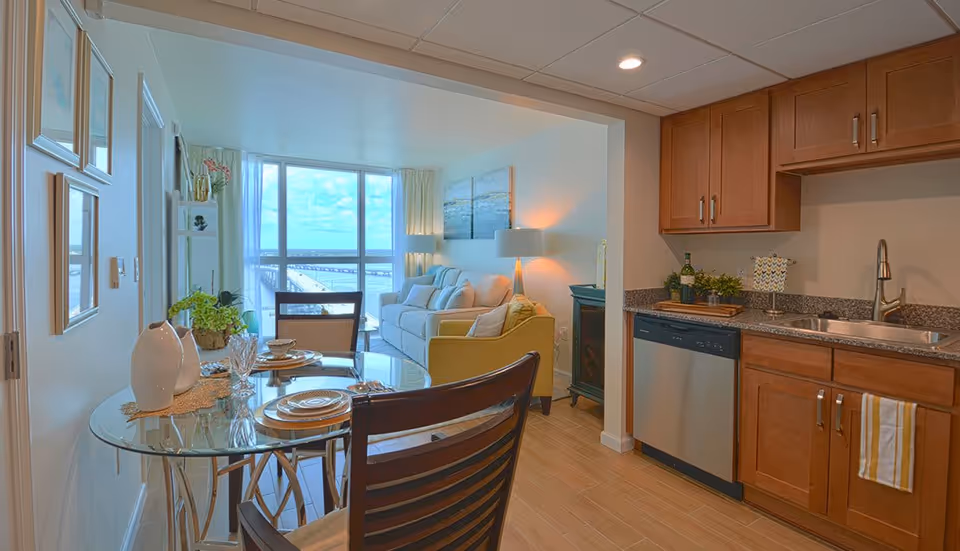 Interior view of a senior living facility apartment featuring a small kitchen with wooden cabinets, a stainless steel dishwasher, and a sink. Adjacent to the kitchen is a dining area with a glass-top table set for two, and a living room with a beige sofa, yellow armchair, and a large window offering a view of a bridge and water outside.